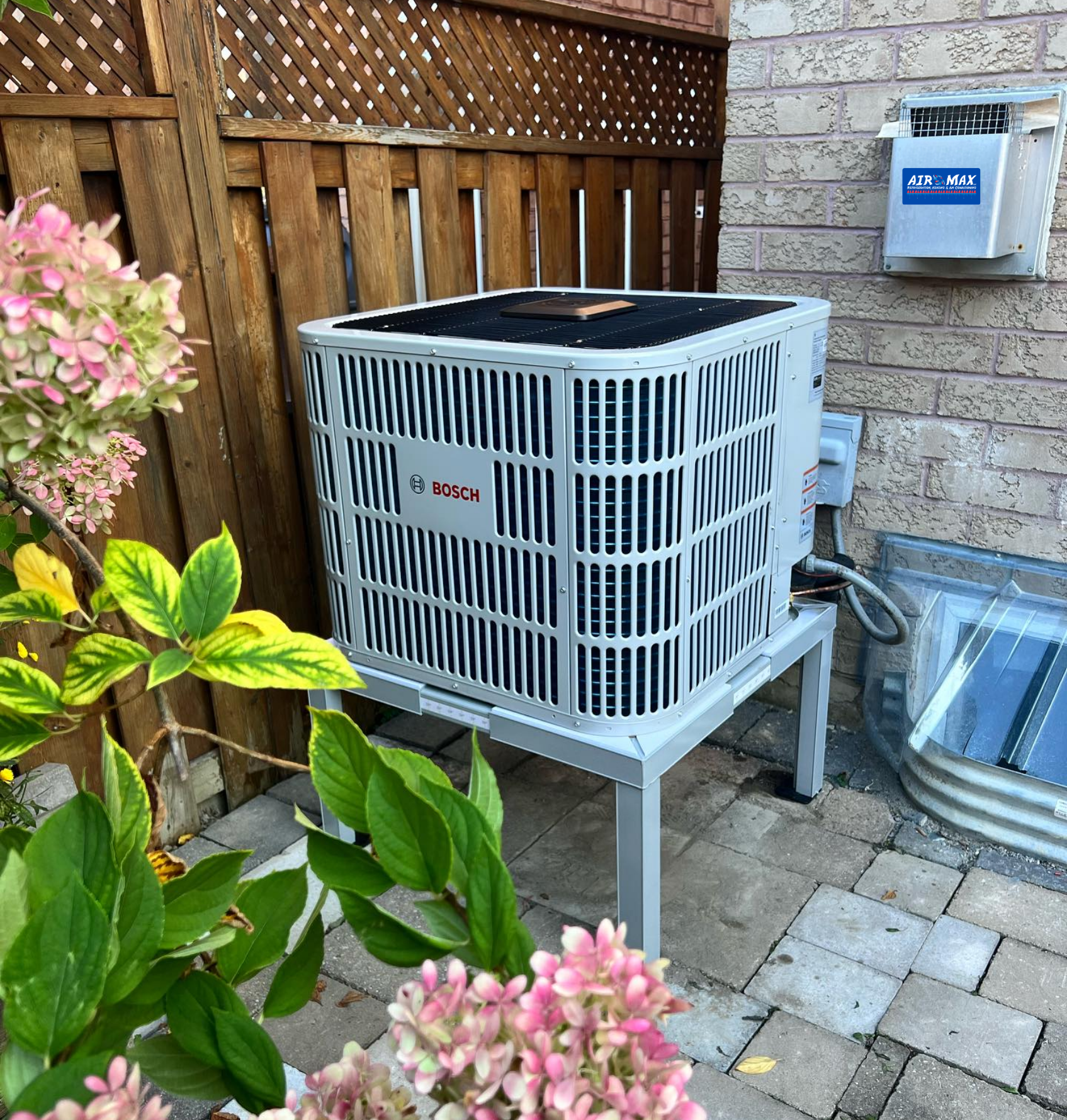 AC unit on a metal stand next to a brick wall and fence, with flowers in the foreground.