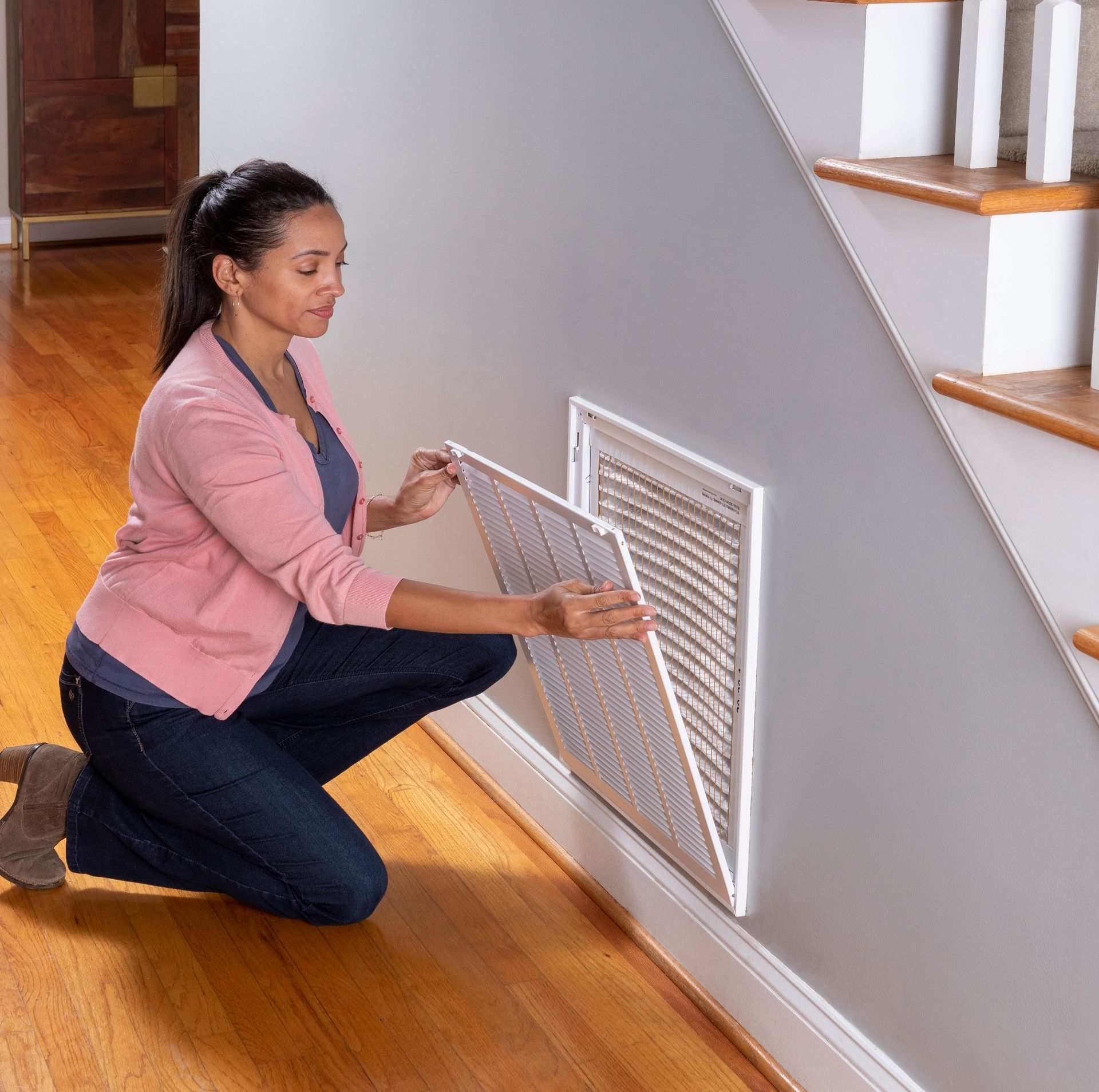 Woman kneels to remove a white vent cover on a gray wall next to wooden stairs.