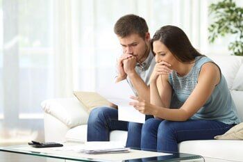 Couple looks at paperwork, concerned expressions. White sofa, living room setting.