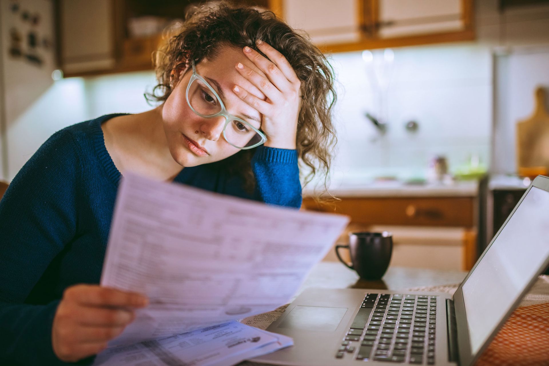 Woman with glasses looks stressed, holding paperwork, next to a laptop and coffee mug.