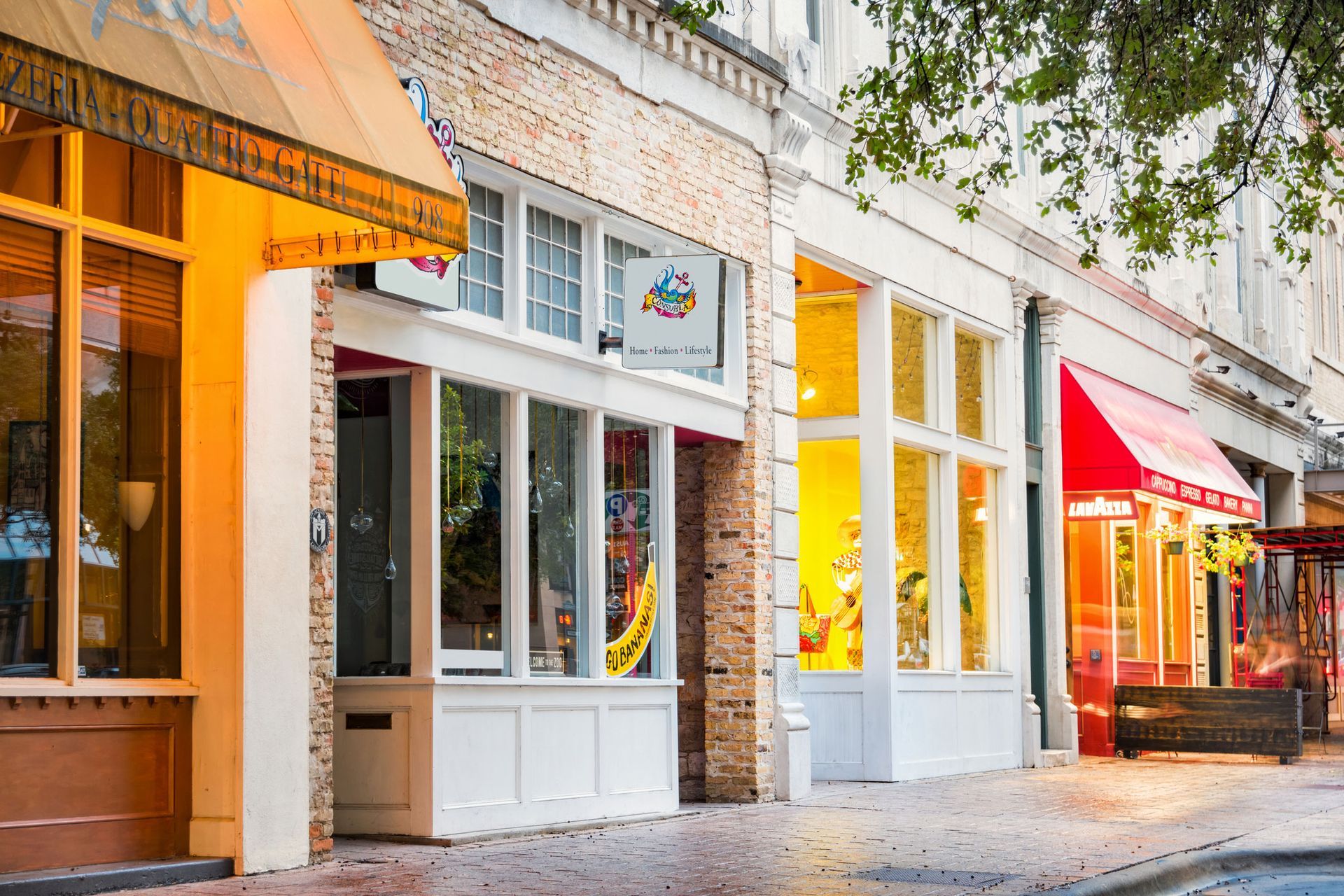 Shops with awnings line a brick-paved street. Warm interior lighting glows through the windows.
