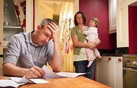 Man stressed, reviewing bills at kitchen table; woman holds child, looking concerned.