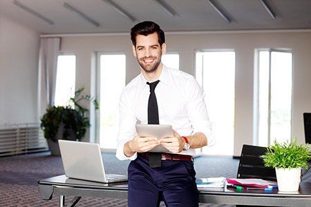 Man in white shirt and tie smiling, holding tablet, standing at a desk with laptop in office setting.