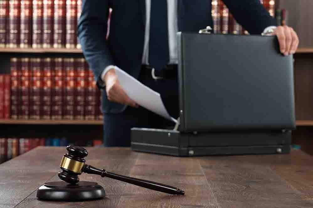 A person in a suit removes papers from a briefcase on a desk next to a gavel and bookshelf.