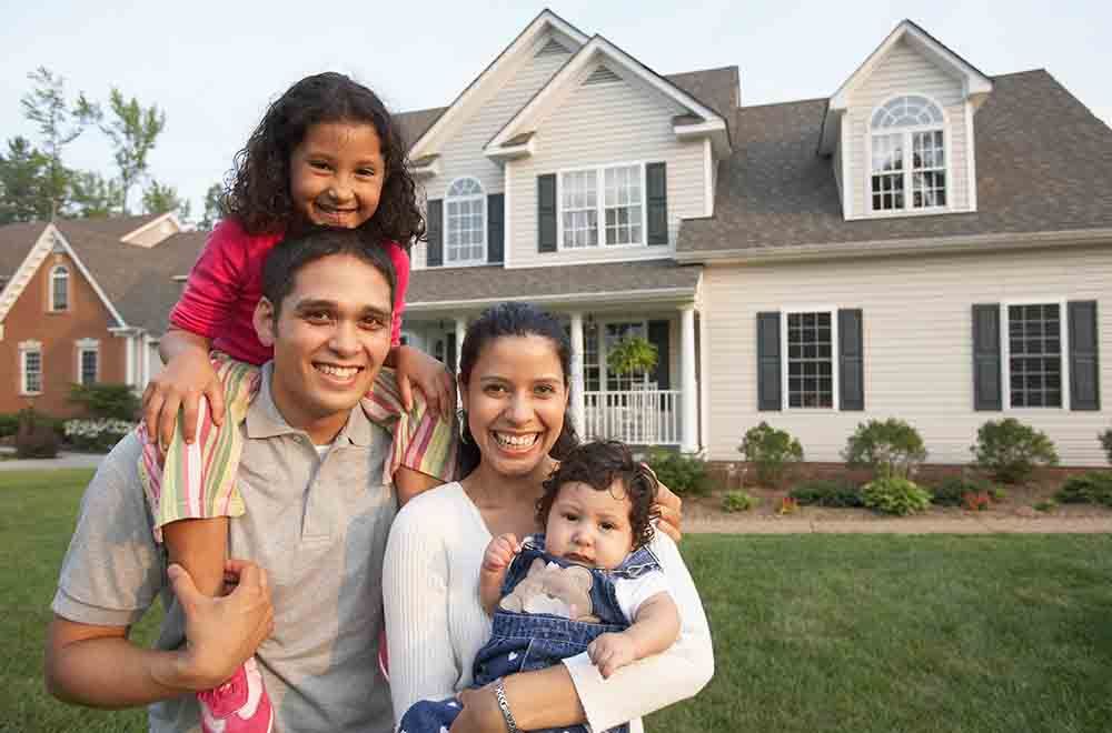 Family of four smiling in front of a house: child on father's shoulders, mother holding baby.