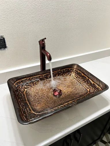 Dark brown ornate glass sink with water running, bronze faucet on white countertop.