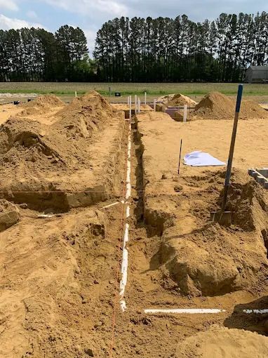 Construction site, trench with white pipes, dirt mounds, shovel, and blueprint.