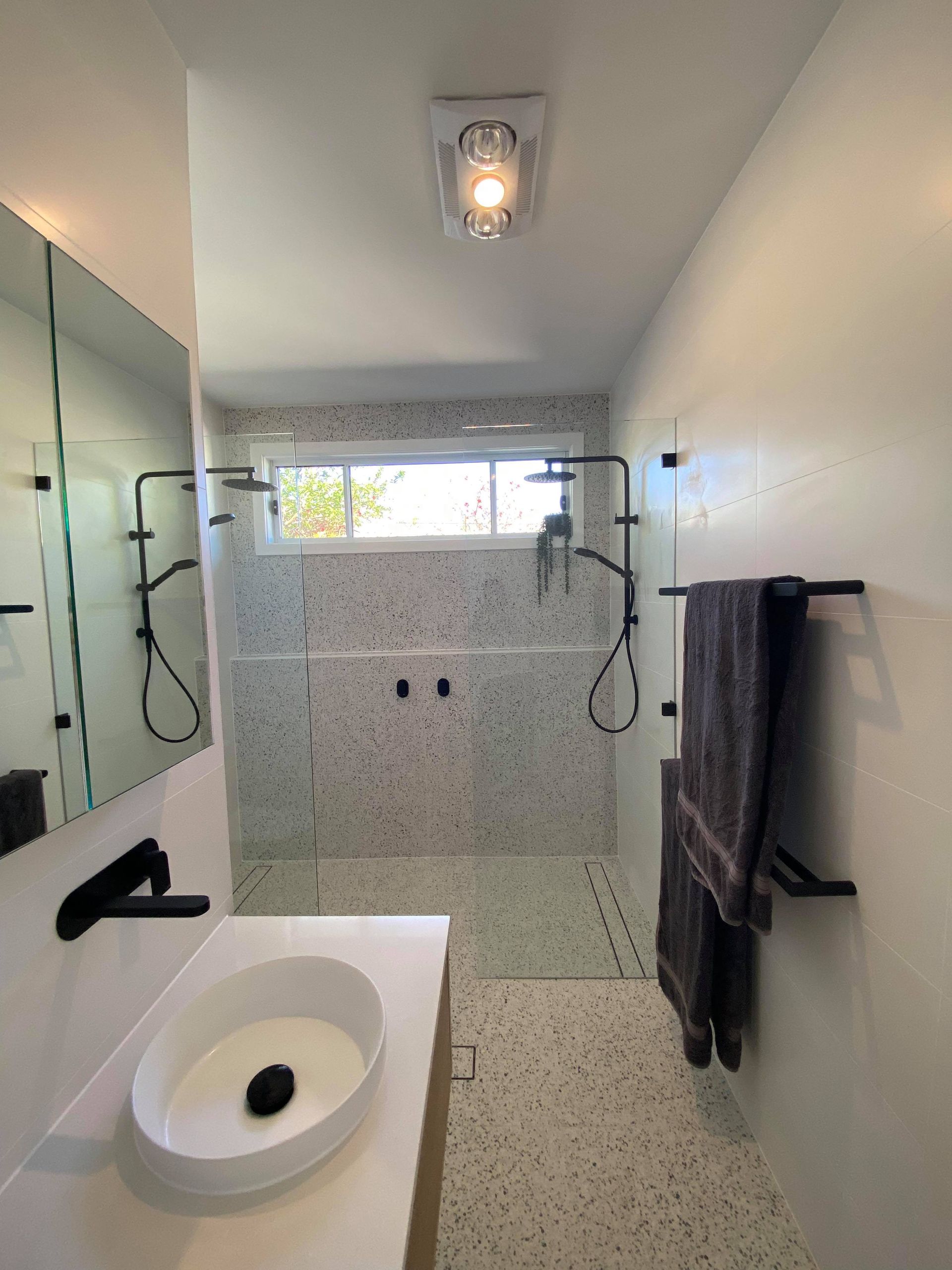 Modern bathroom with double showerheads, speckled tile, sink, mirror, and towel bars