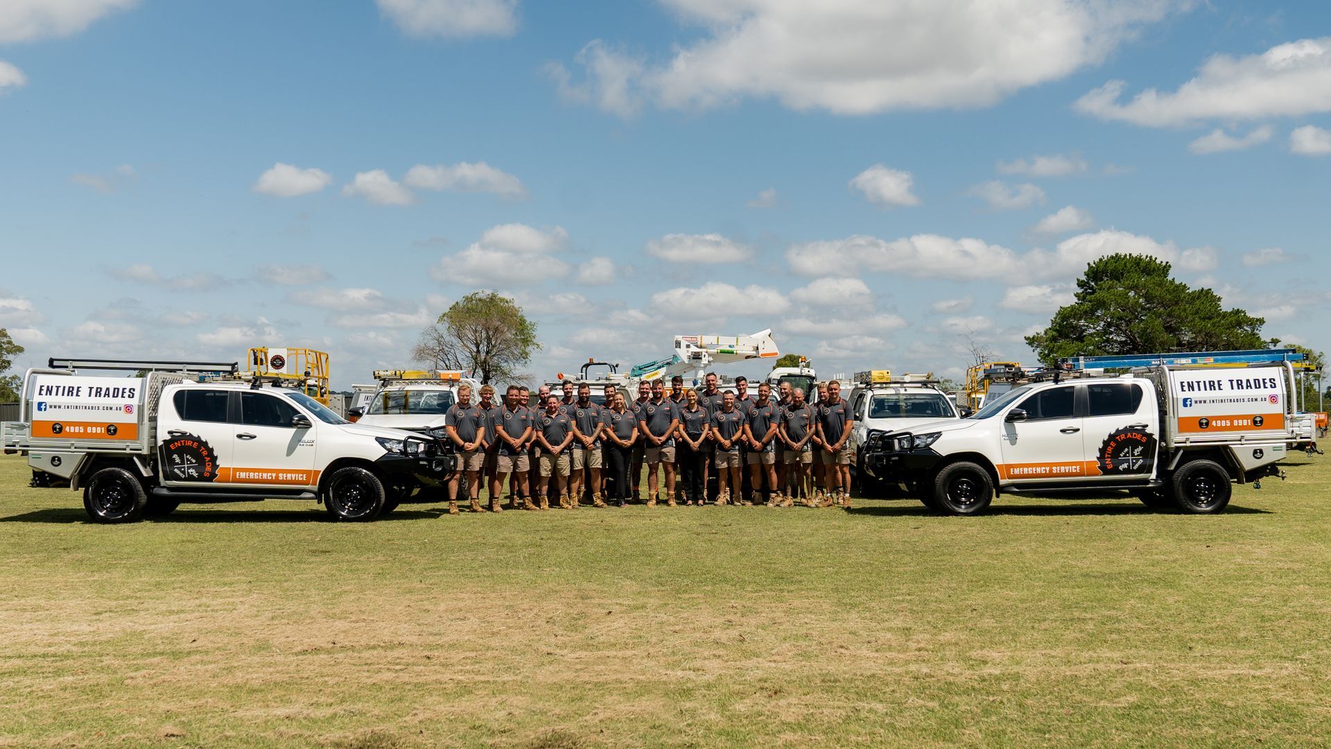 Group of volunteers posing in a field beside utility trucks and equipment under a partly cloudy sky