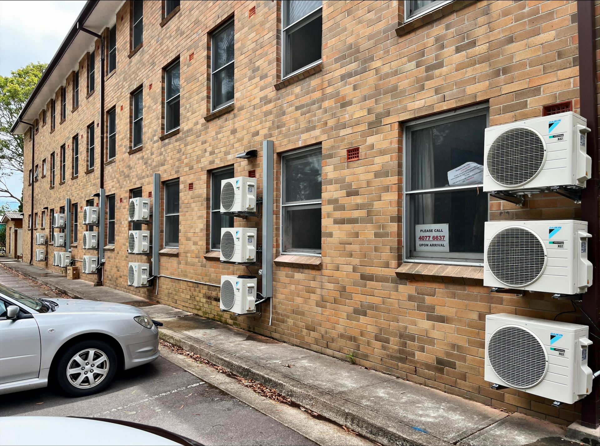Six white outdoor air conditioning units mounted on a brick wall beside windows.