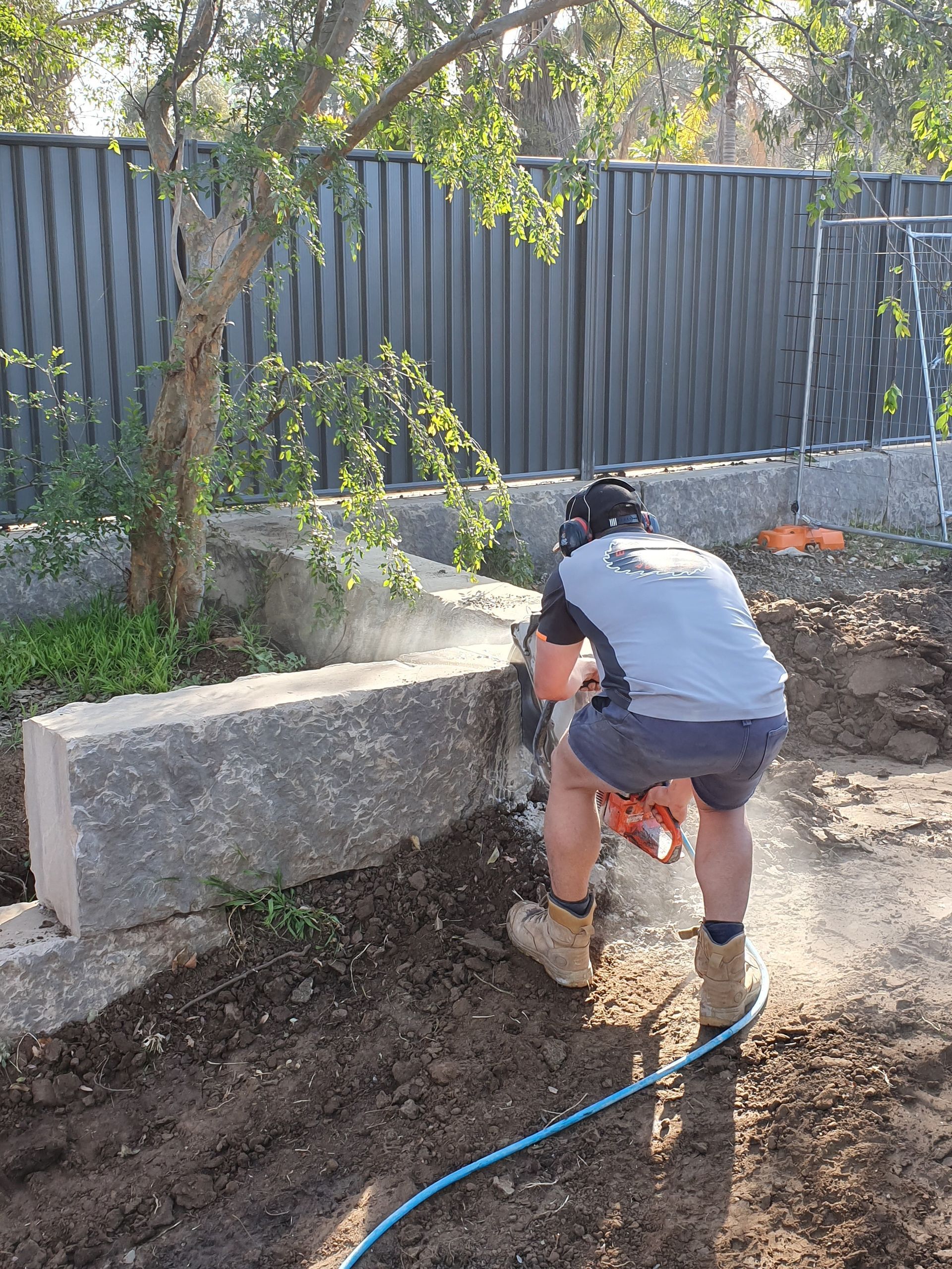 Person kneeling in a muddy yard, working near a concrete block wall and blue hose.