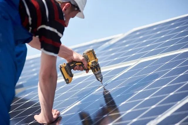 Worker installing solar panels with a drill on a rooftop under a blue sky