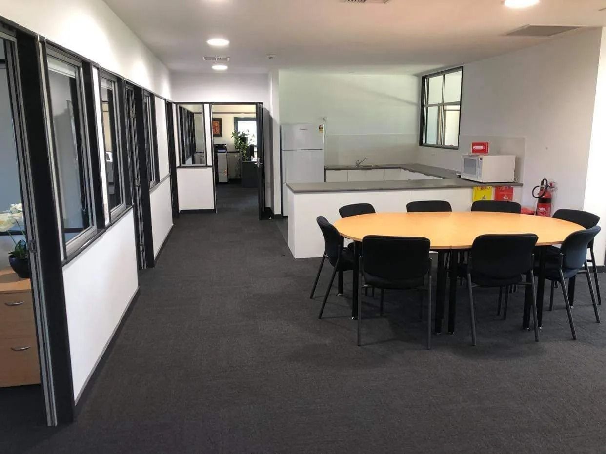 Office meeting area with a round table, black chairs, and a hallway beside windows and white walls