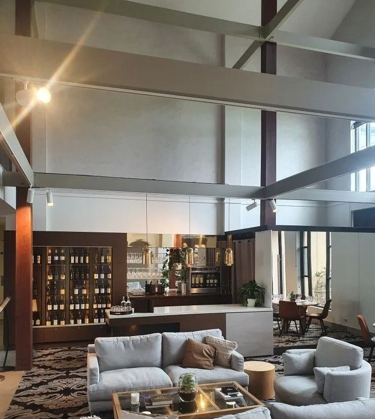 Modern living room with tall windows, exposed beams, gray seating, and a bar area in the background