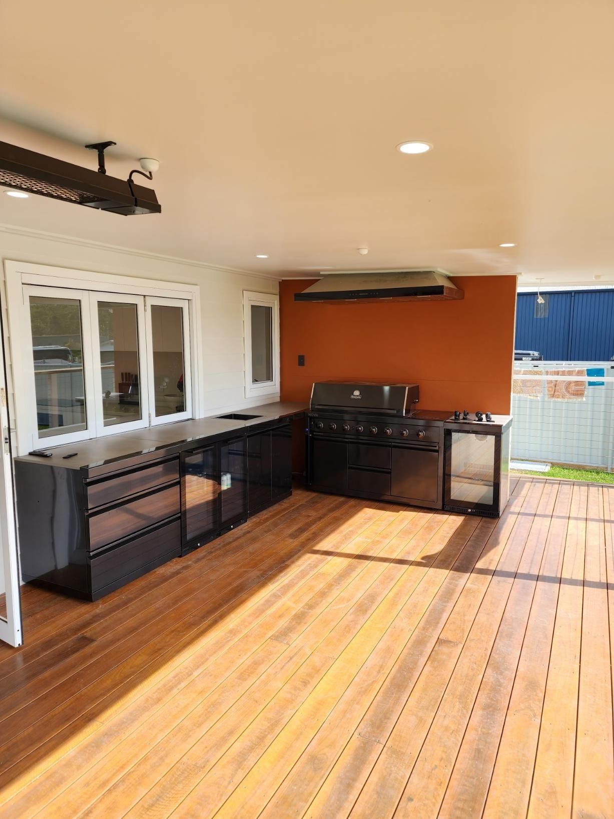 Outdoor kitchen with black cabinets and stove on a sunlit wooden deck, beside a white window and orange wall