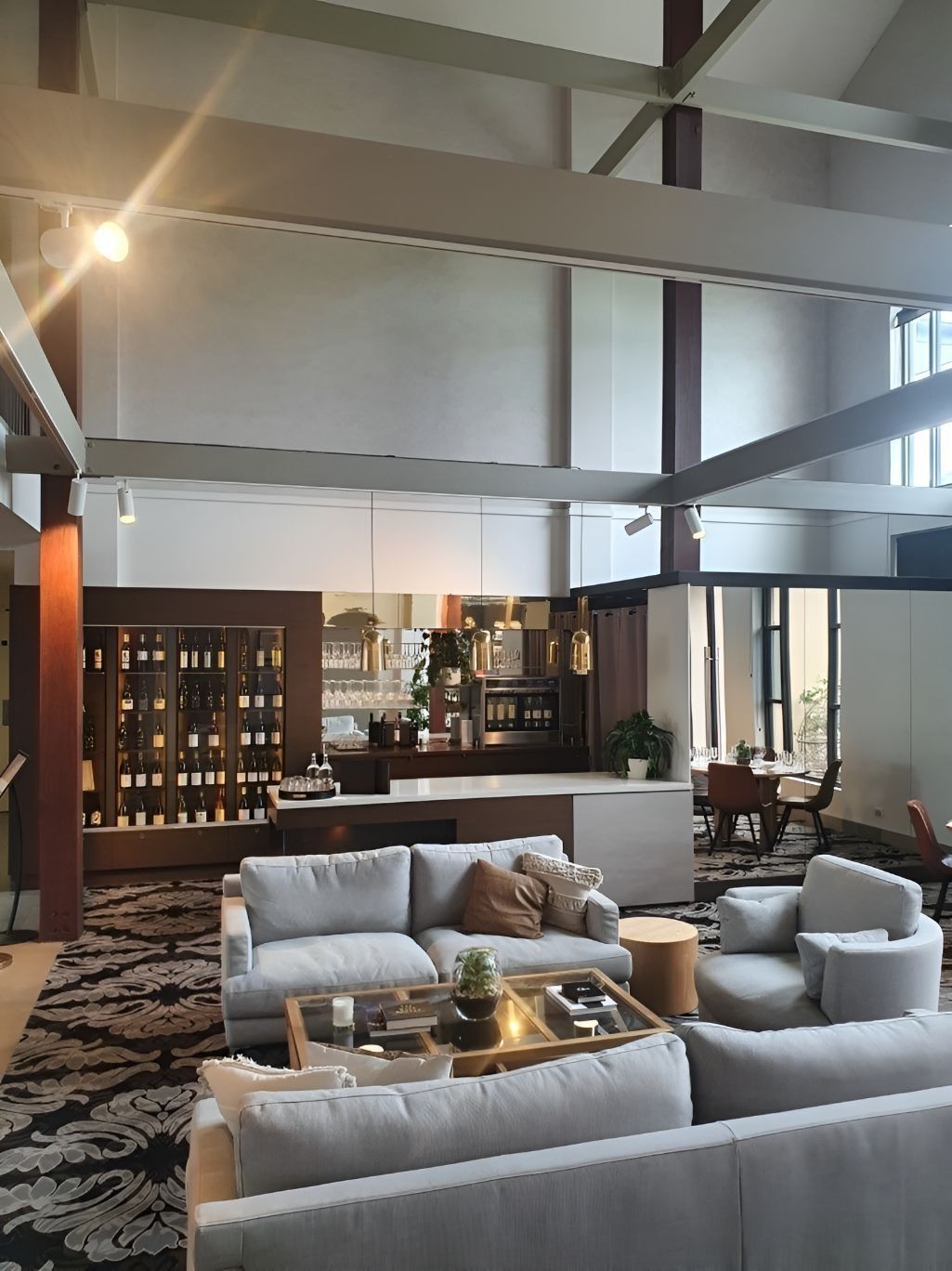 Modern lounge with gray sofas, patterned rug, wood bar, and tall vaulted ceiling with exposed beams