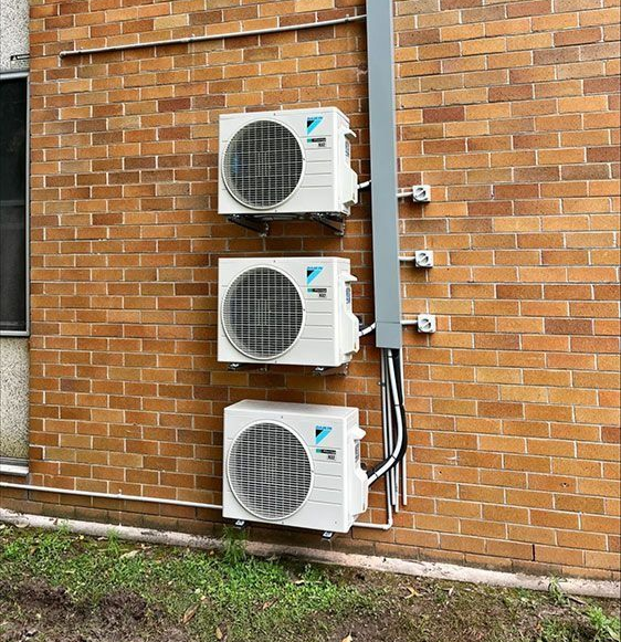 Three wall-mounted outdoor AC units on a brick building with exposed pipes and wiring.