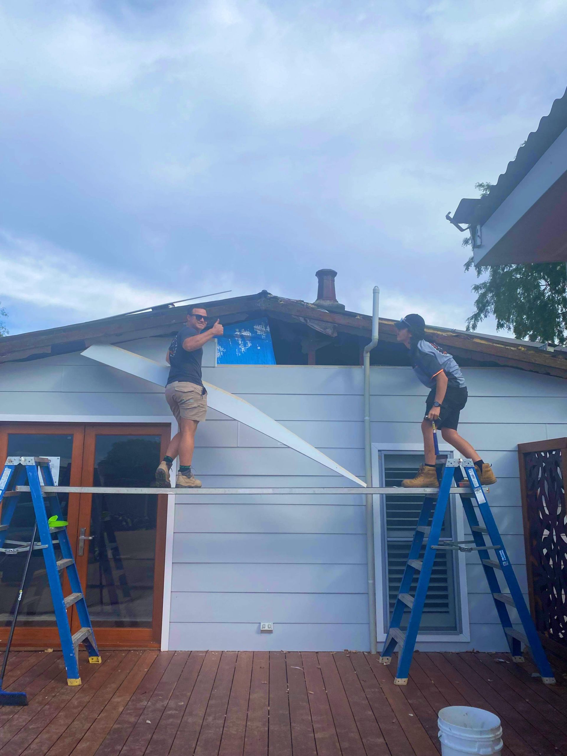 Two workers installing siding on a house, standing on ladders under a cloudy sky.