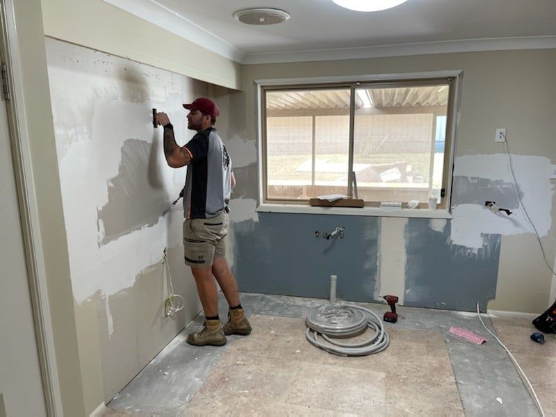 Worker mudding drywall in a room with a window and renovation tools on the floor