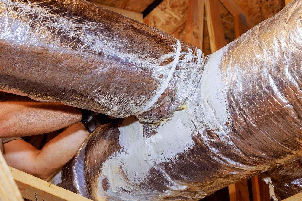 Foil-wrapped HVAC ductwork in an attic with a hand working between the ducts