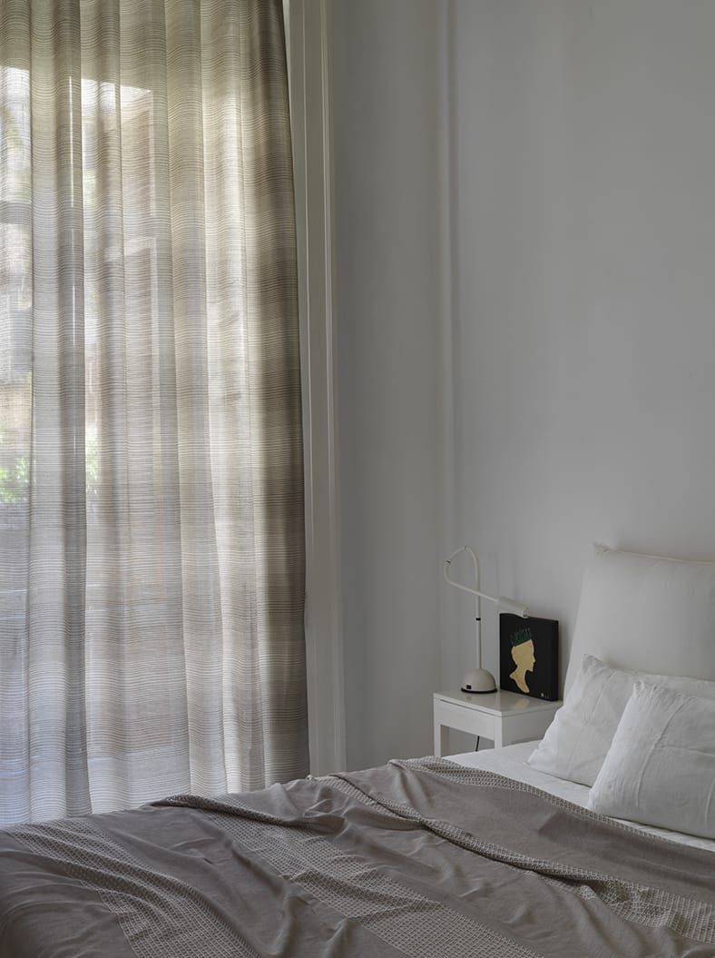 Camera da letto con tende trasparenti, pareti bianche, letto con cuscini e comodino.