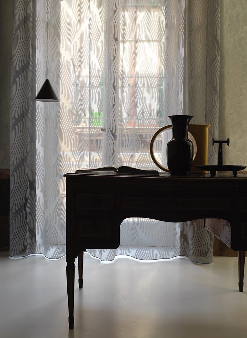 Scrivania con silhouette, vaso e lampada sospesa, davanti a una finestra con tende trasparenti a fantasia.