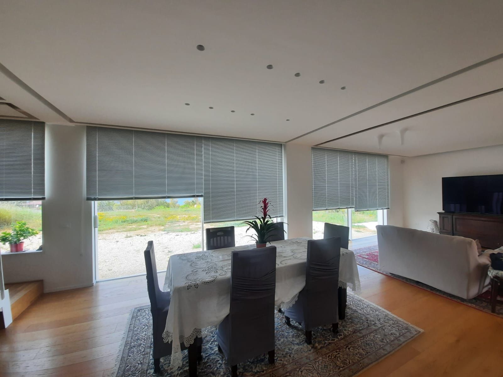 Sala da pranzo con tavolo apparecchiato, ampie finestre con tende e zona soggiorno.