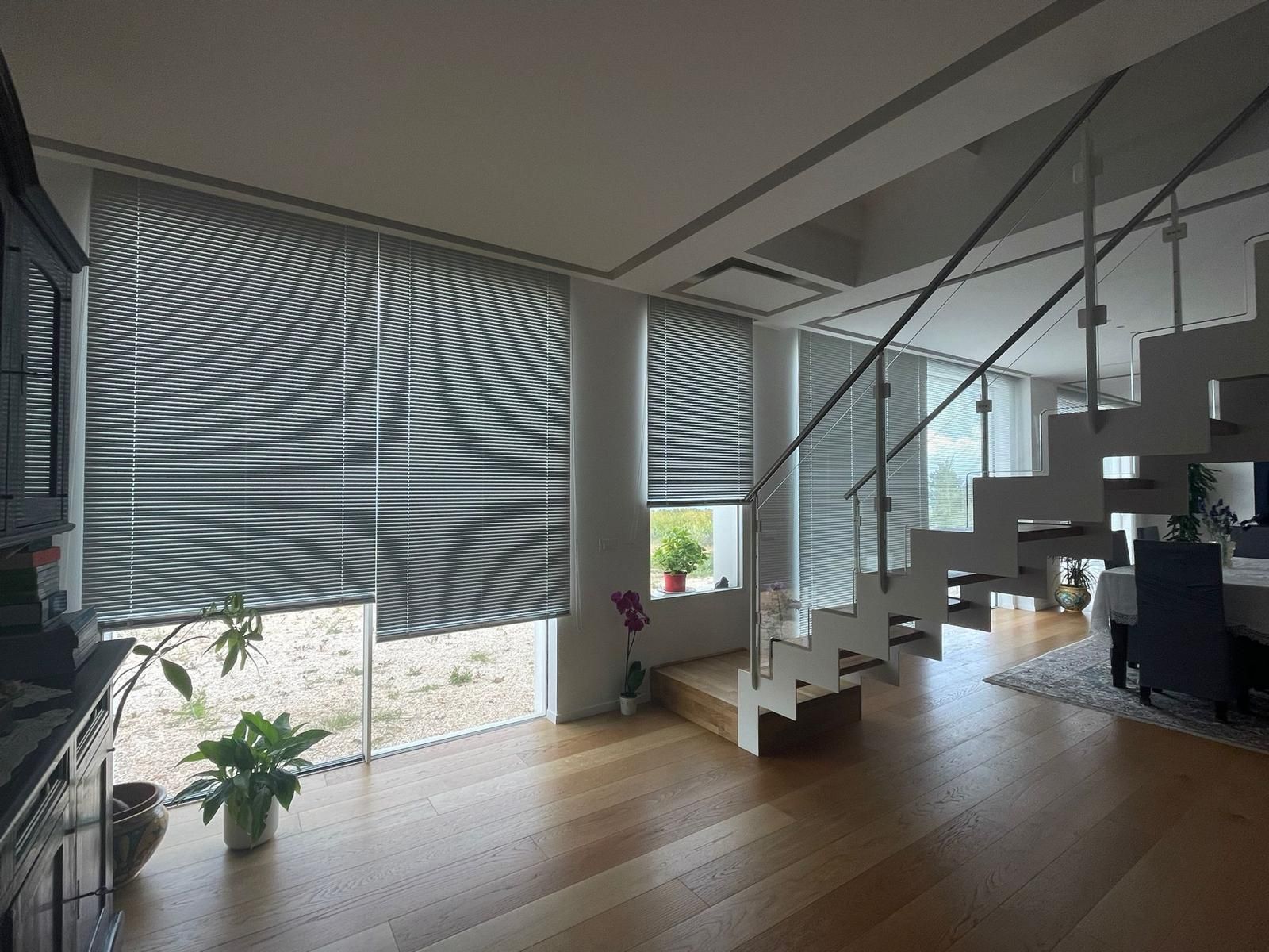 Vista interna di una casa moderna con ampie finestre coperte da persiane e una scala. Pavimenti in legno.