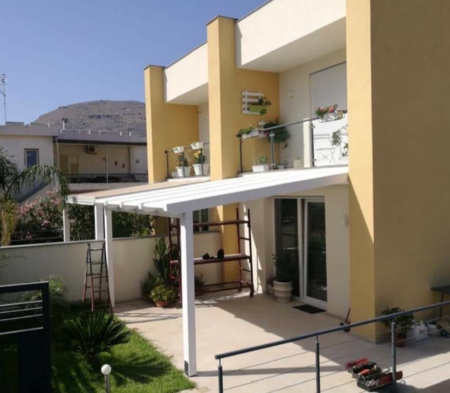 Un pergolato bianco ombreggia il patio di un edificio a due piani con pareti gialle e un balcone con piante.