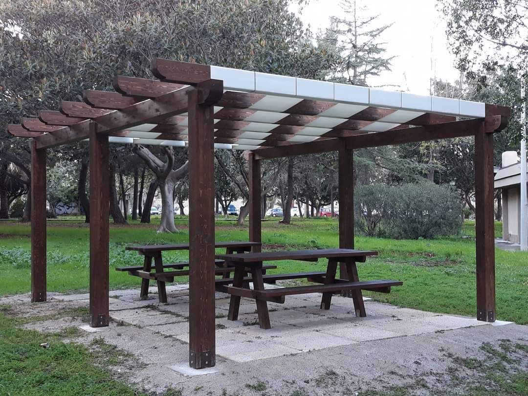 Pergola in legno con tavoli da picnic in un parco, ricoperta da pannelli bianchi alternati.