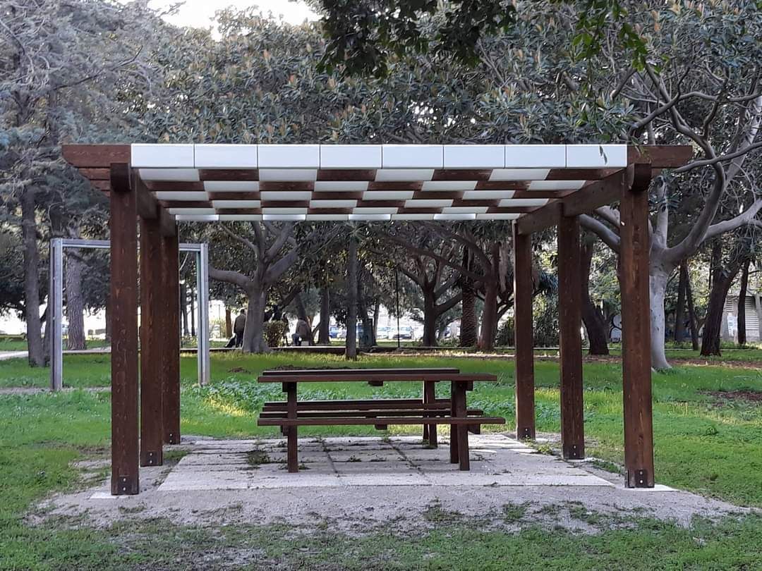 Tavolo da picnic sotto un pergolato in legno in un parco; alberi sullo sfondo. Tetto a scacchiera.
