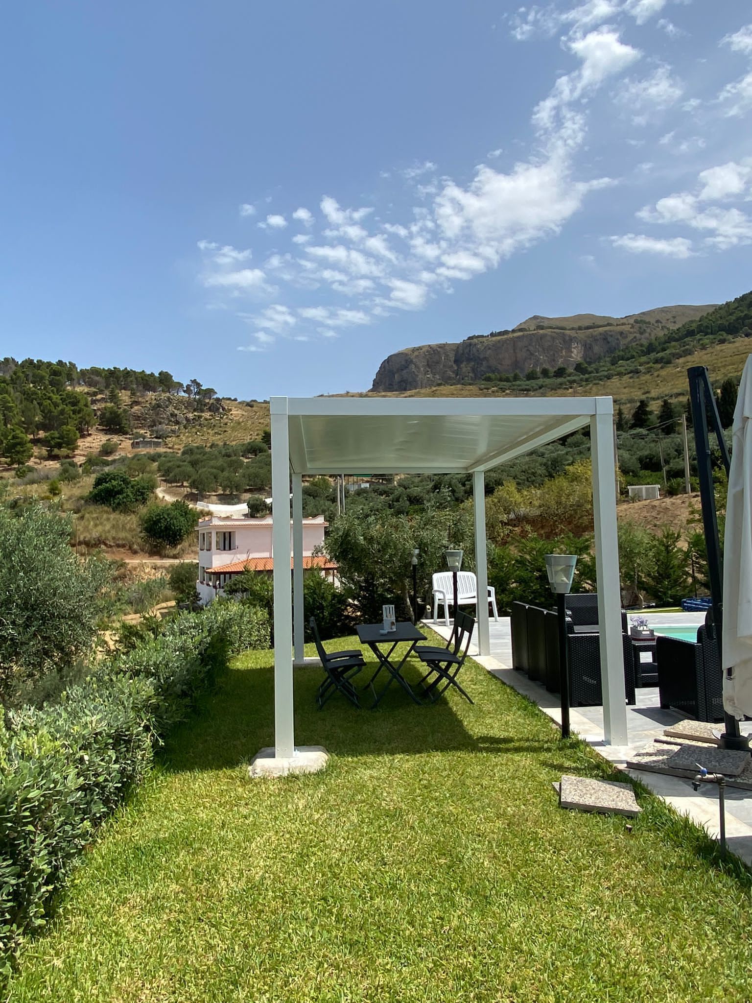 Pergola bianca sopra un tavolo e delle sedie in un cortile erboso, con una montagna sullo sfondo.