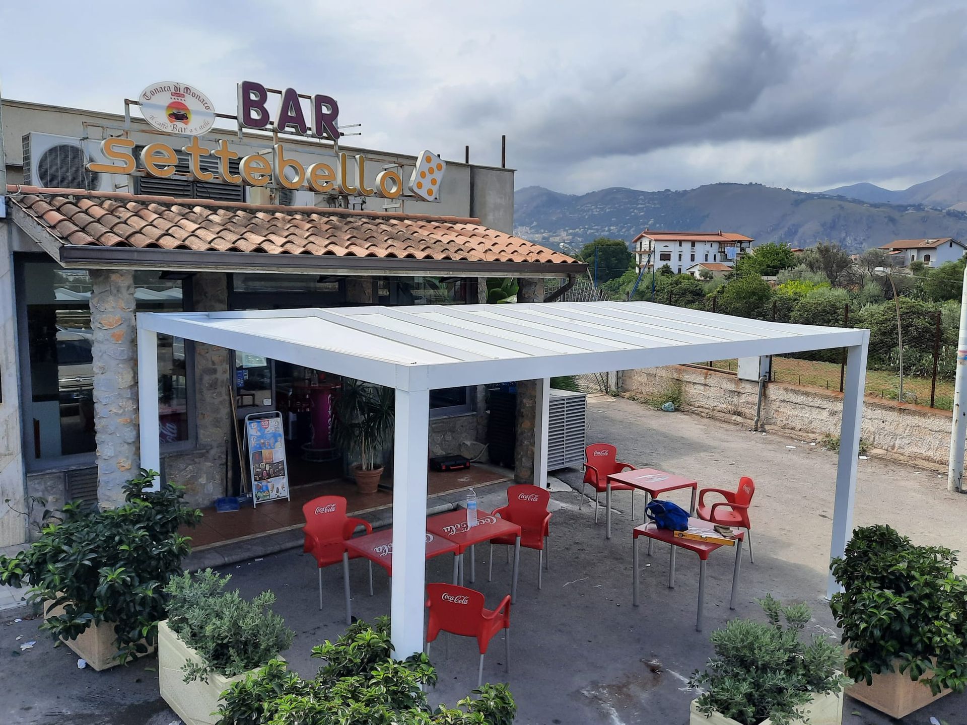 Bar all'aperto con pergolato bianco, tavoli rossi, sedie e insegna del bar.