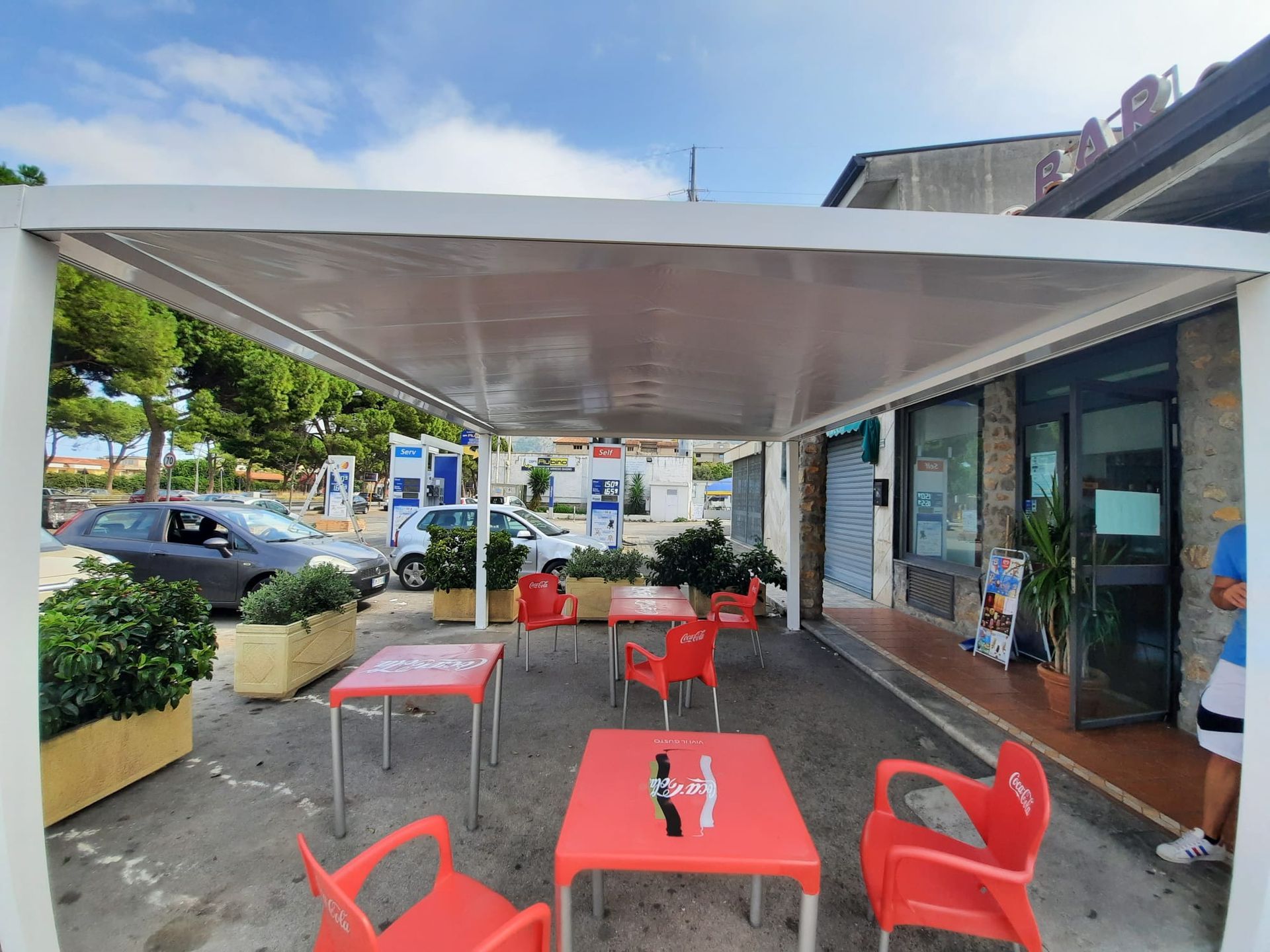 Bar all'aperto con tavoli e sedie rossi sotto una tettoia bianca. Sullo sfondo si vedono automobili e un edificio.