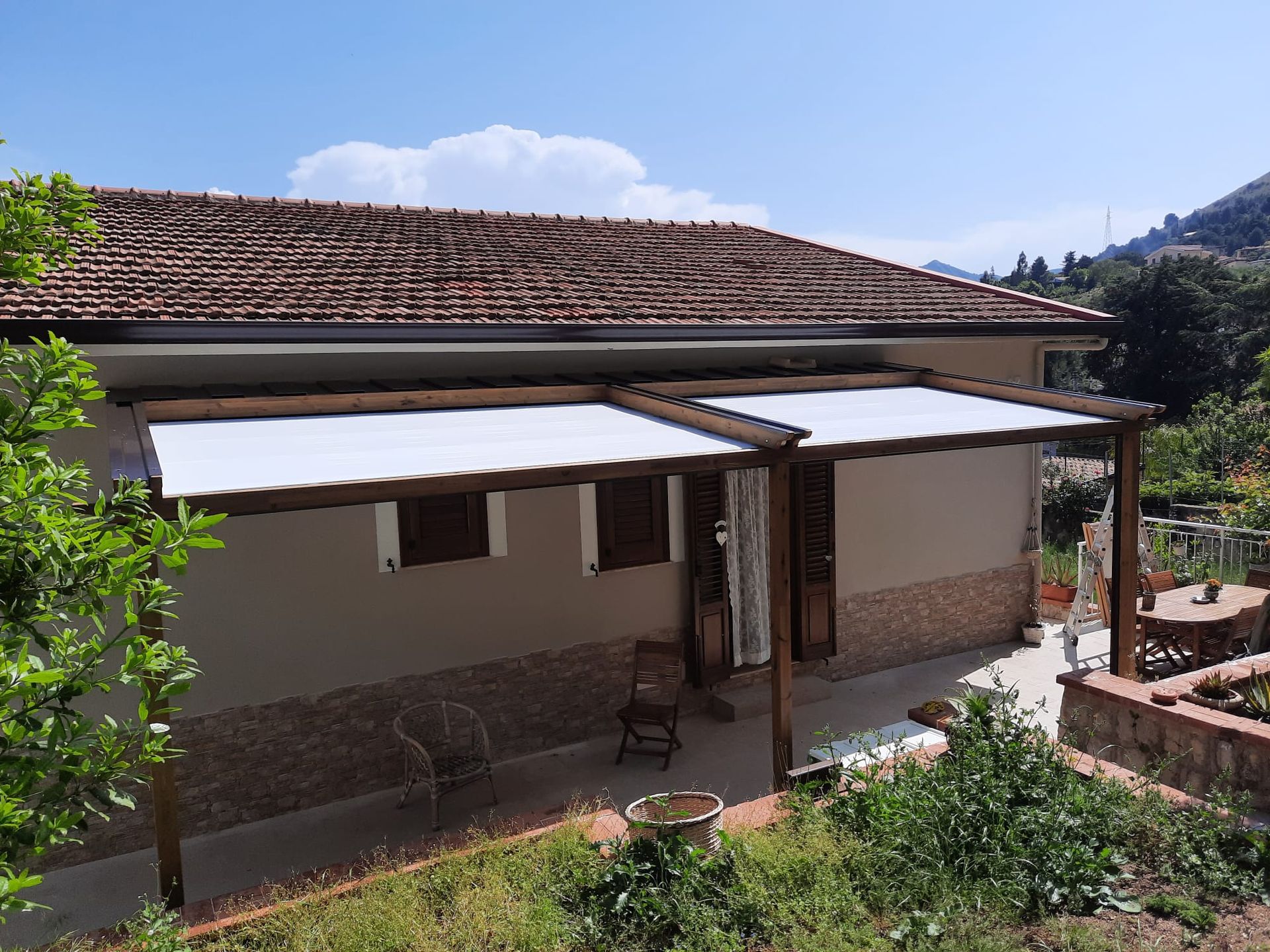 Casa con tetto marrone e pergolato in legno annesso con tettoia bianca.