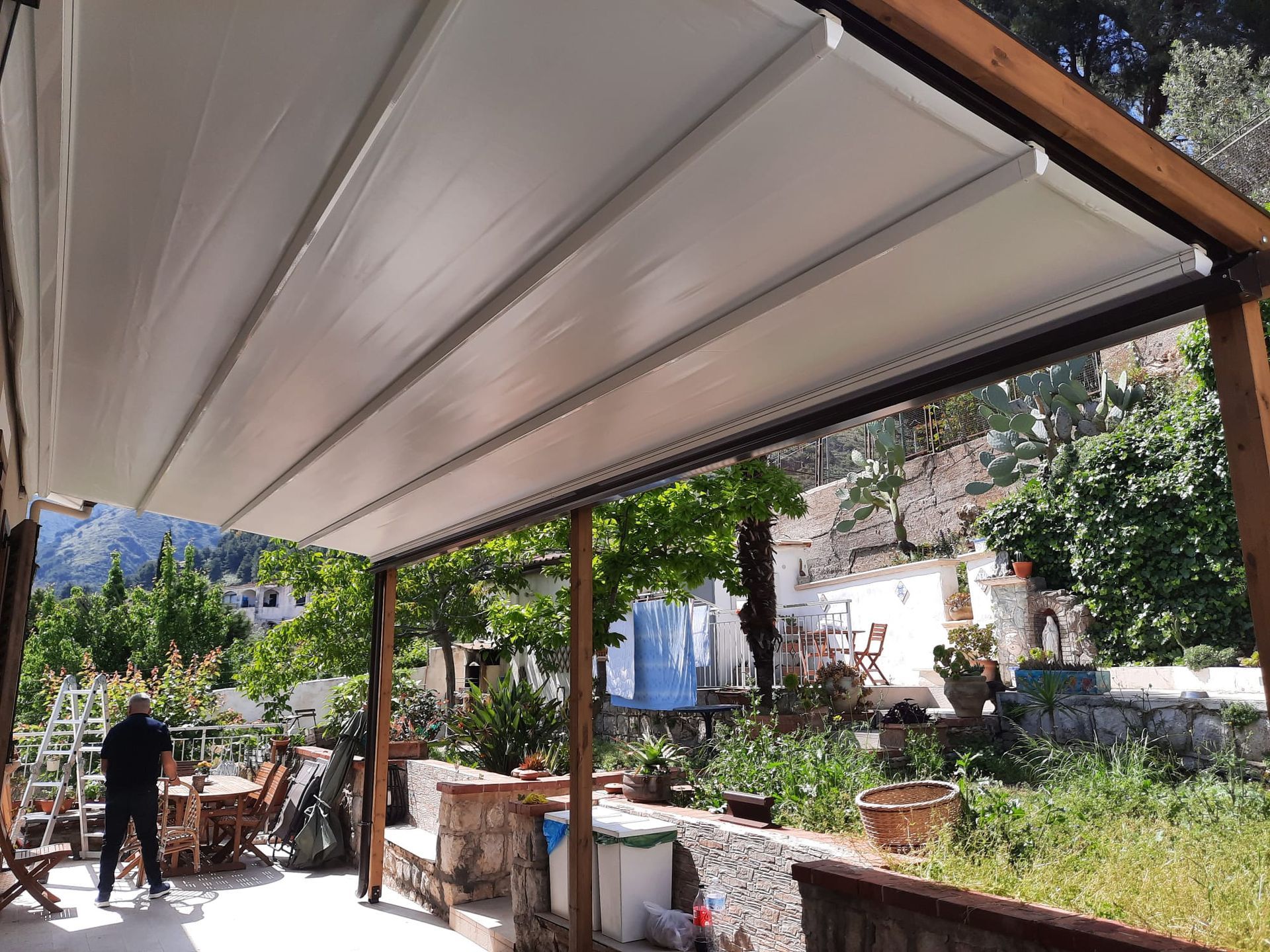 Patio coperto con tettoia bianca e supporti in legno, con vista su un cortile verde.