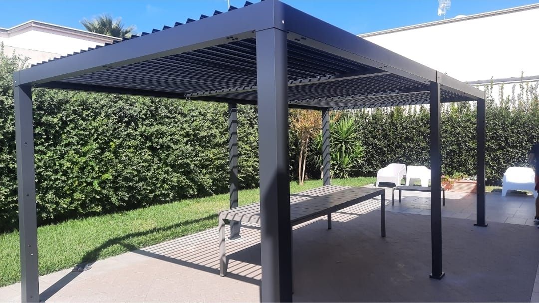 Pergola nera sopra un patio con tavolo e sedie, immersa nel verde. Giornata di sole.