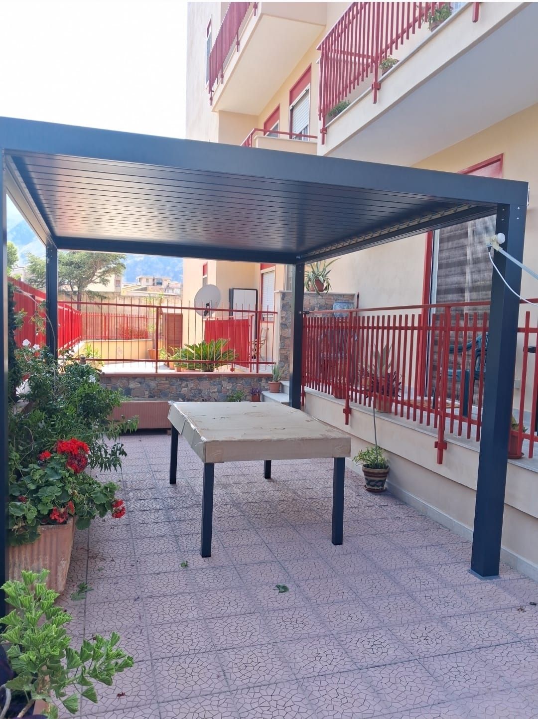 Pergola nera sopra un tavolo da giardino. Sullo sfondo, ringhiere rosse e un edificio. Piante in vaso in primo piano.