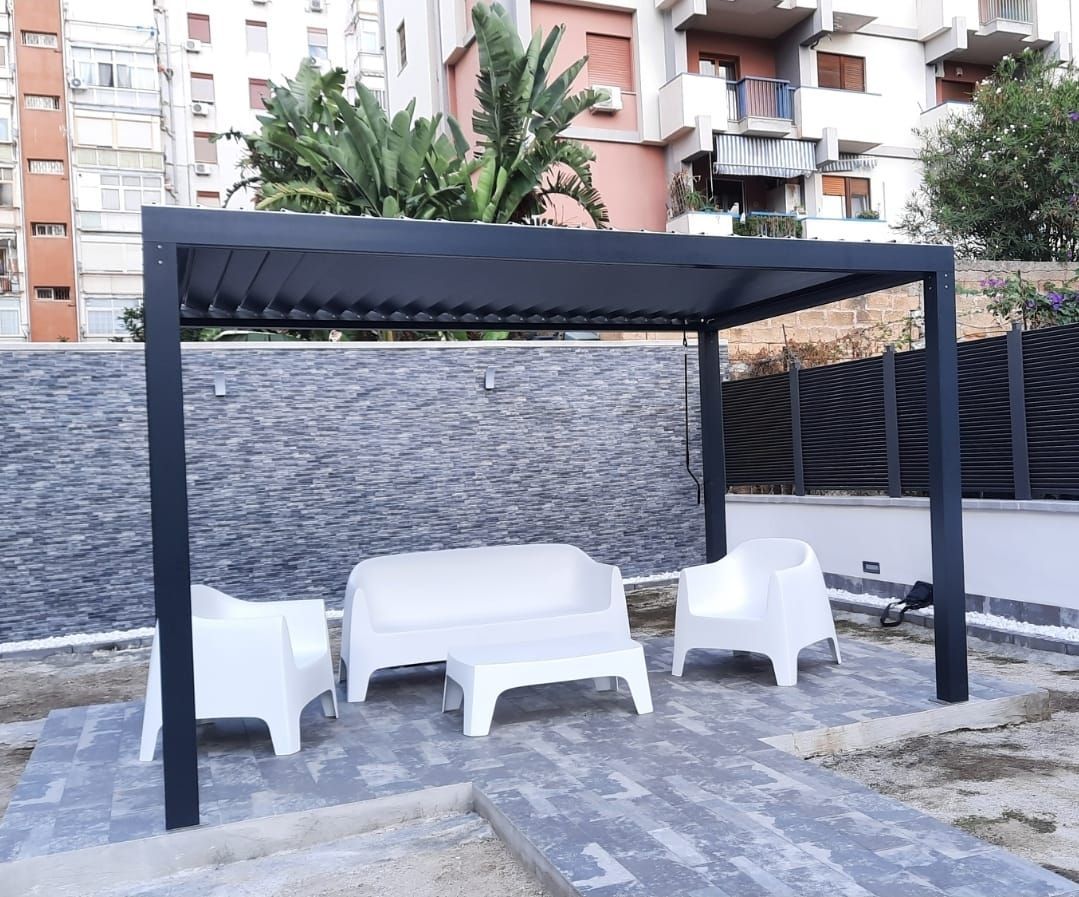 Pergola con zona salotto: struttura grigio scuro, mobili bianchi, sfondo con muro in pietra, ambiente esterno.