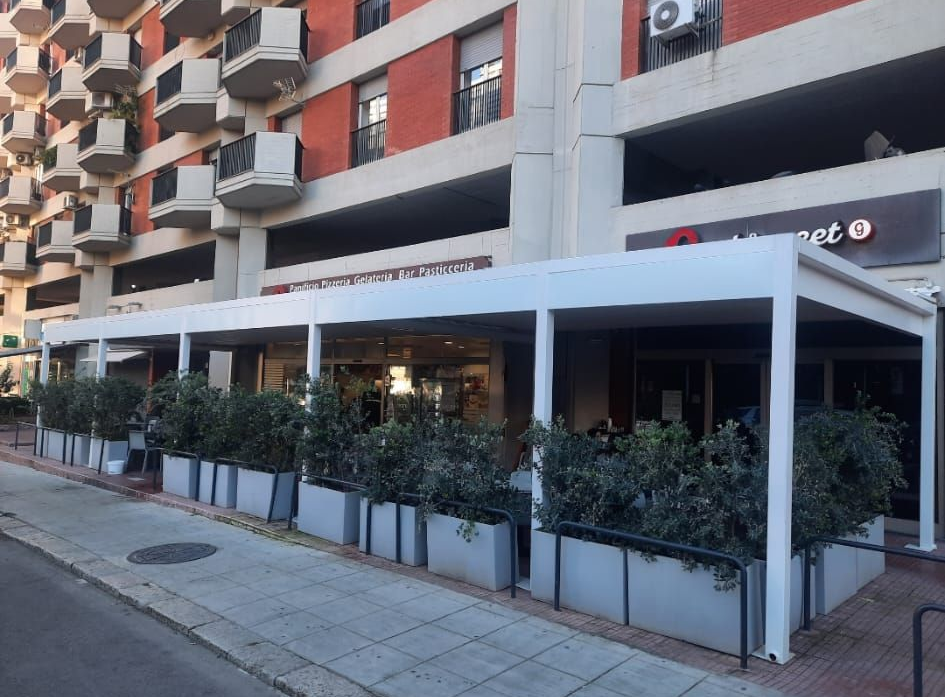 Ristorante con pergolato bianco, posti a sedere all'aperto e fioriere con cespugli verdi. Un edificio sullo sfondo.