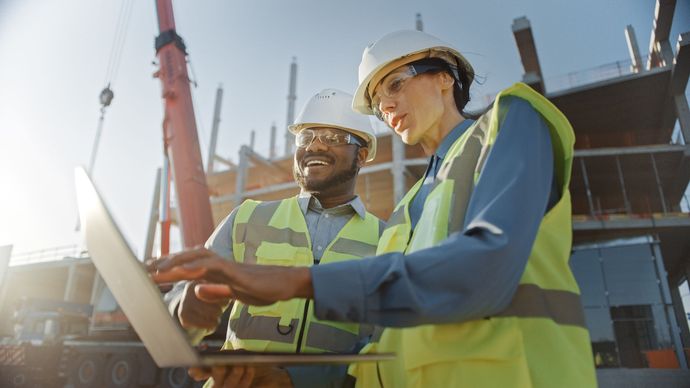 Two construction workers in hard hats and safety vests reviewing plans at a building site