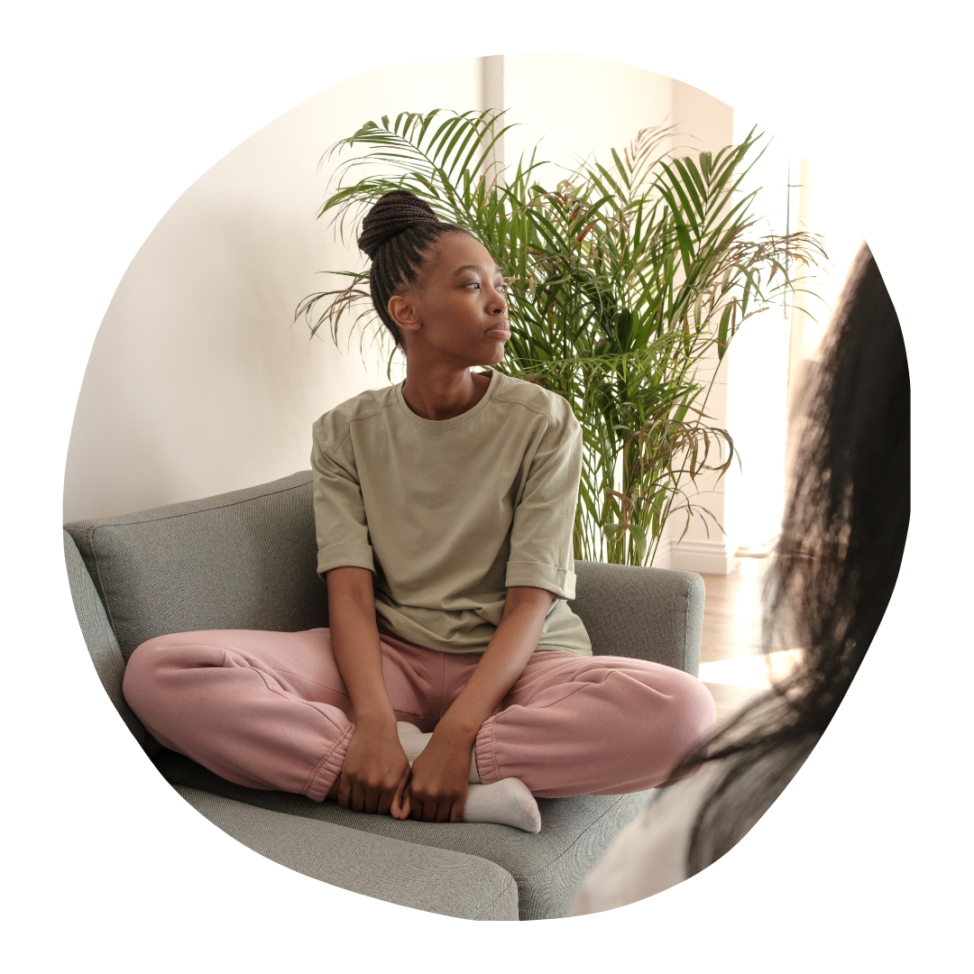 Person sitting cross-legged on a gray couch in a bright room with a large potted plant behind them.