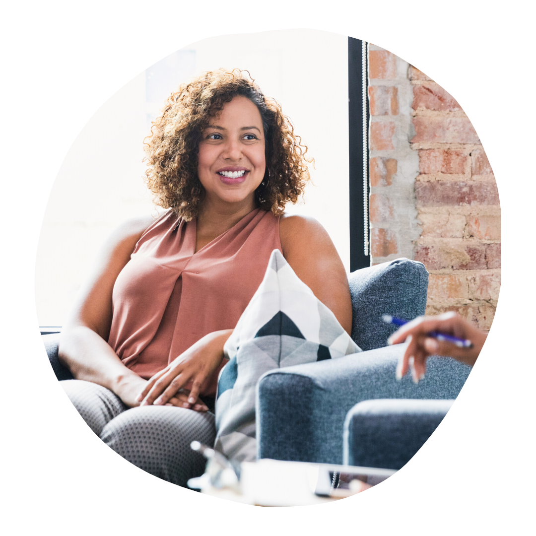Smiling woman in a peach top seated in a bright living room with blue chairs and a brick wall