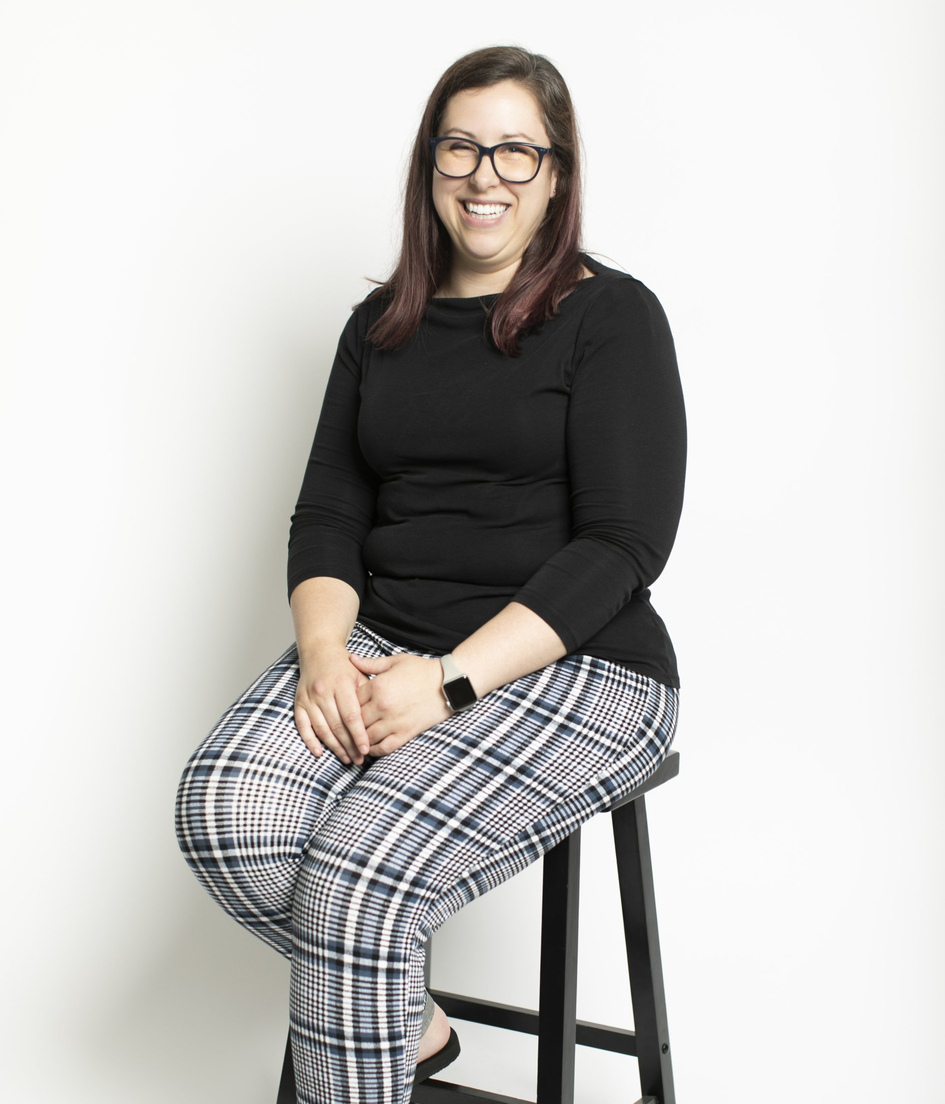 Smiling person in black top and plaid pants seated on a stool against a white background