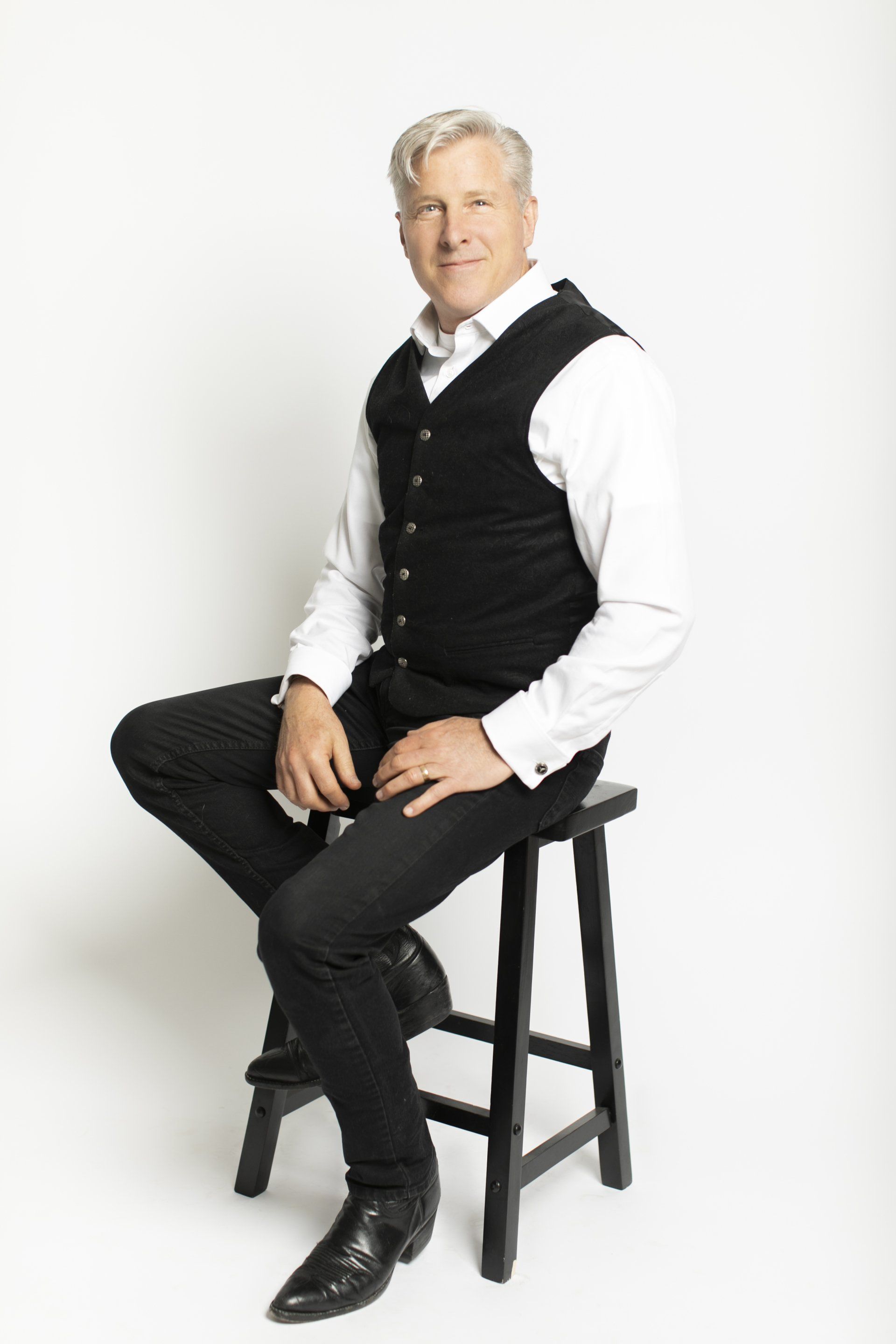 Man seated on a black stool, wearing a white shirt, black vest, and black pants against a white background