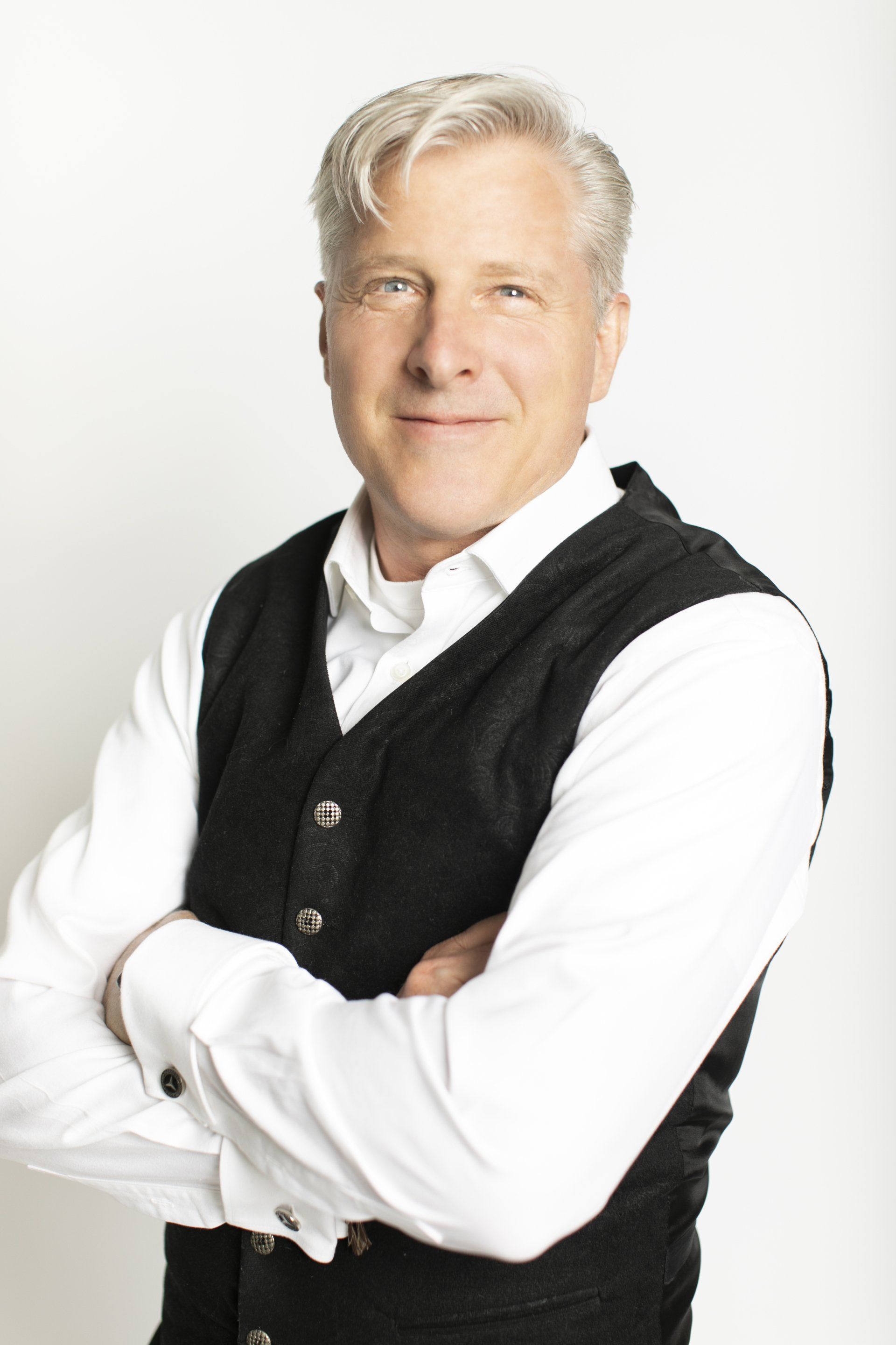 Portrait of a smiling man in a white shirt and black vest, standing with arms crossed against a light background