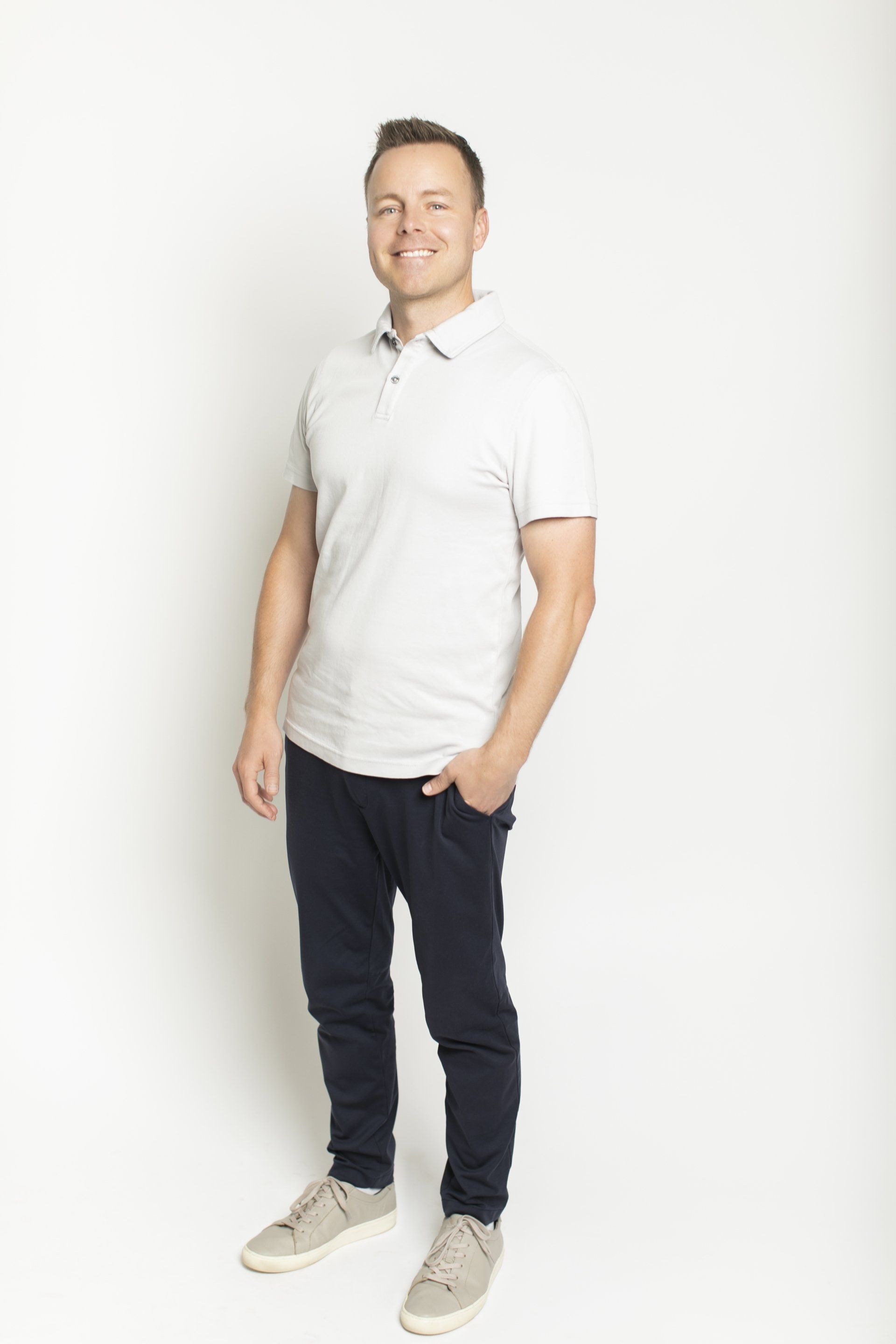 Man standing in a white polo, dark pants, and white sneakers against a plain white background