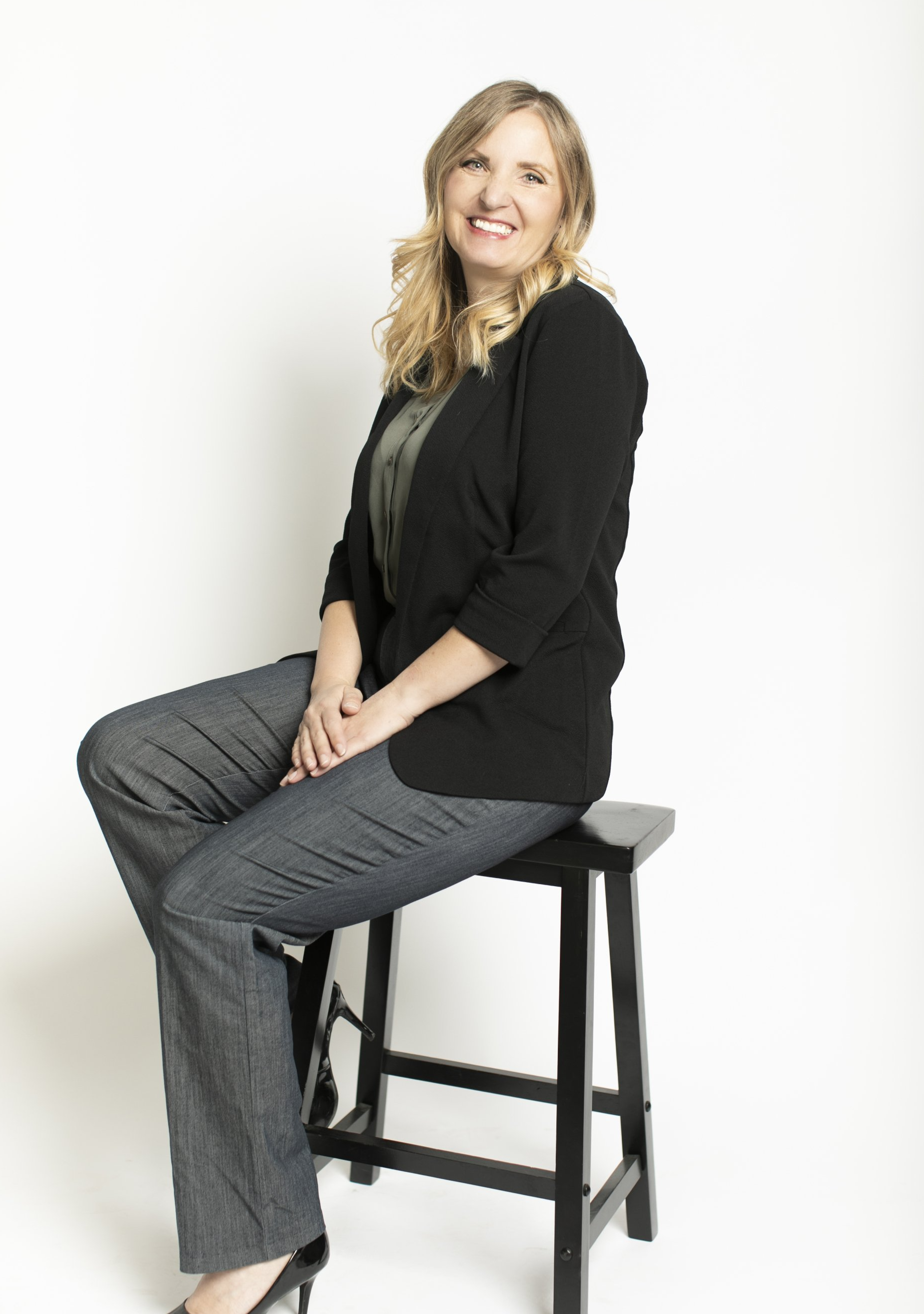 Woman seated on a stool, smiling, in a black blazer and gray pants against a white background.
