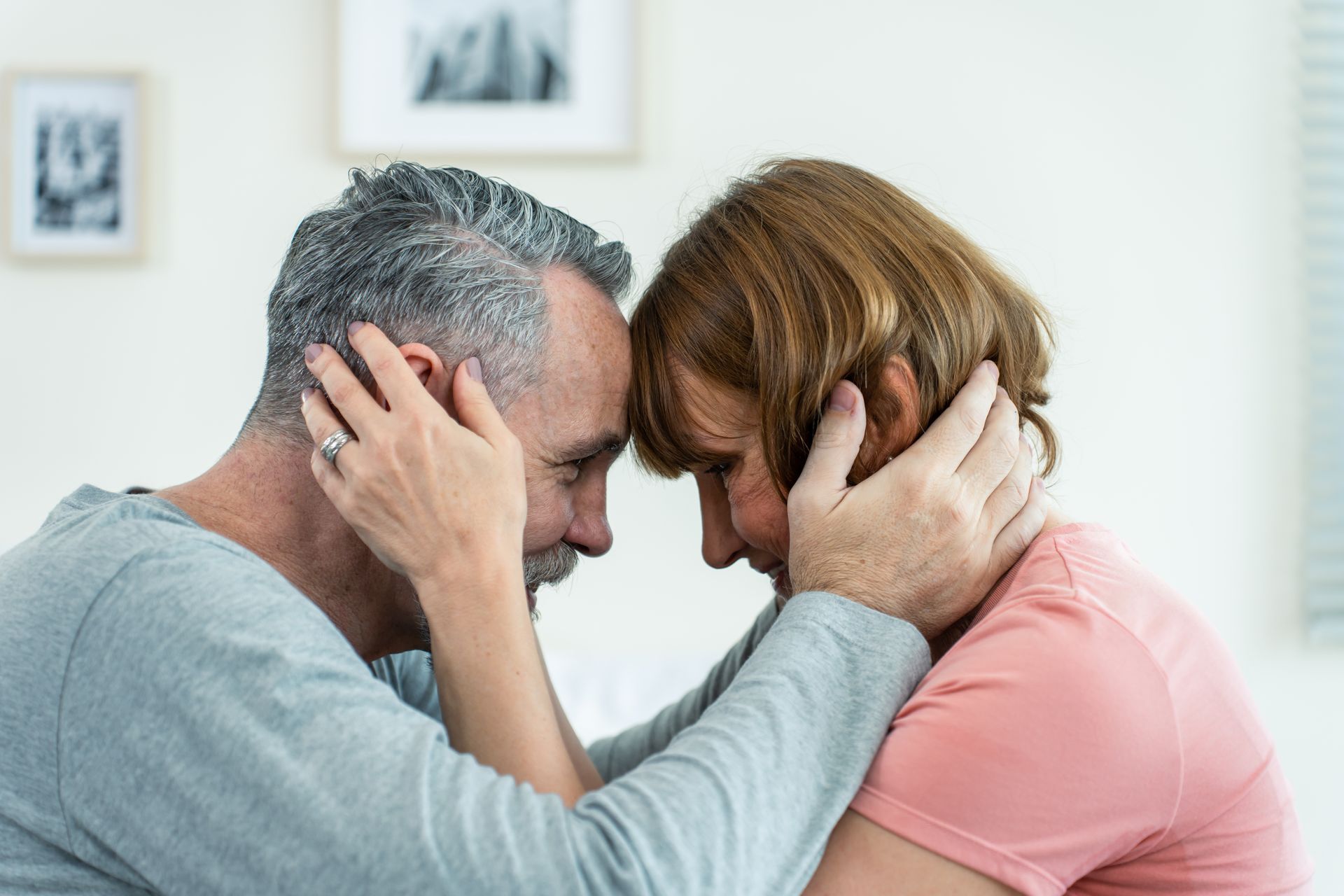 Two adults sit close, foreheads touching and holding each other’s faces in a bright room.