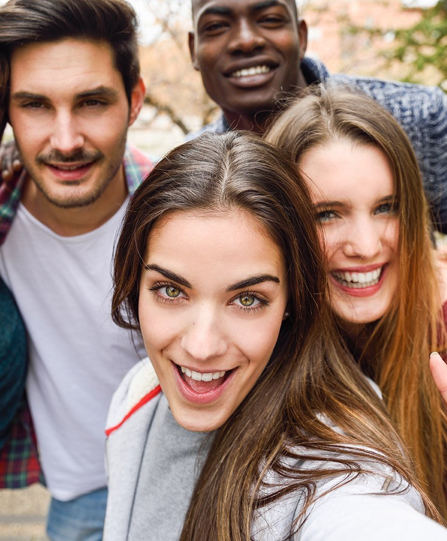 Four friends smiling closely outdoors, with trees blurred in the background.